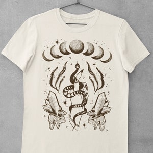 Pu&ograve; includere: Una maglietta beige con una stampa grafica marrone di una mano che tiene un serpente, cristalli e un design a mezzaluna.