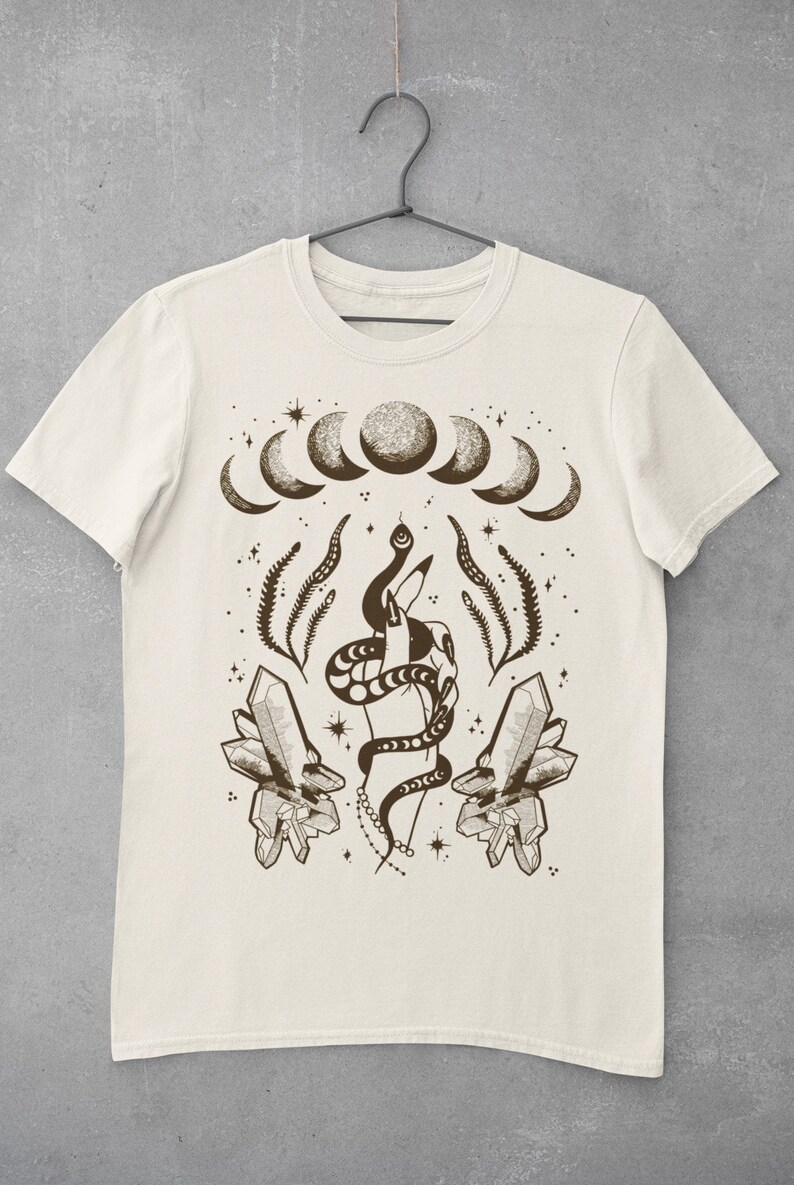 Pu&ograve; includere: Una maglietta beige con una stampa grafica marrone di una mano che tiene un serpente, cristalli e un design a mezzaluna.