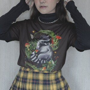 May include: A black t-shirt with a raccoon illustration surrounded by leaves, mushrooms, and stars. The shirt is worn over a black turtleneck and paired with a yellow and black plaid skirt.