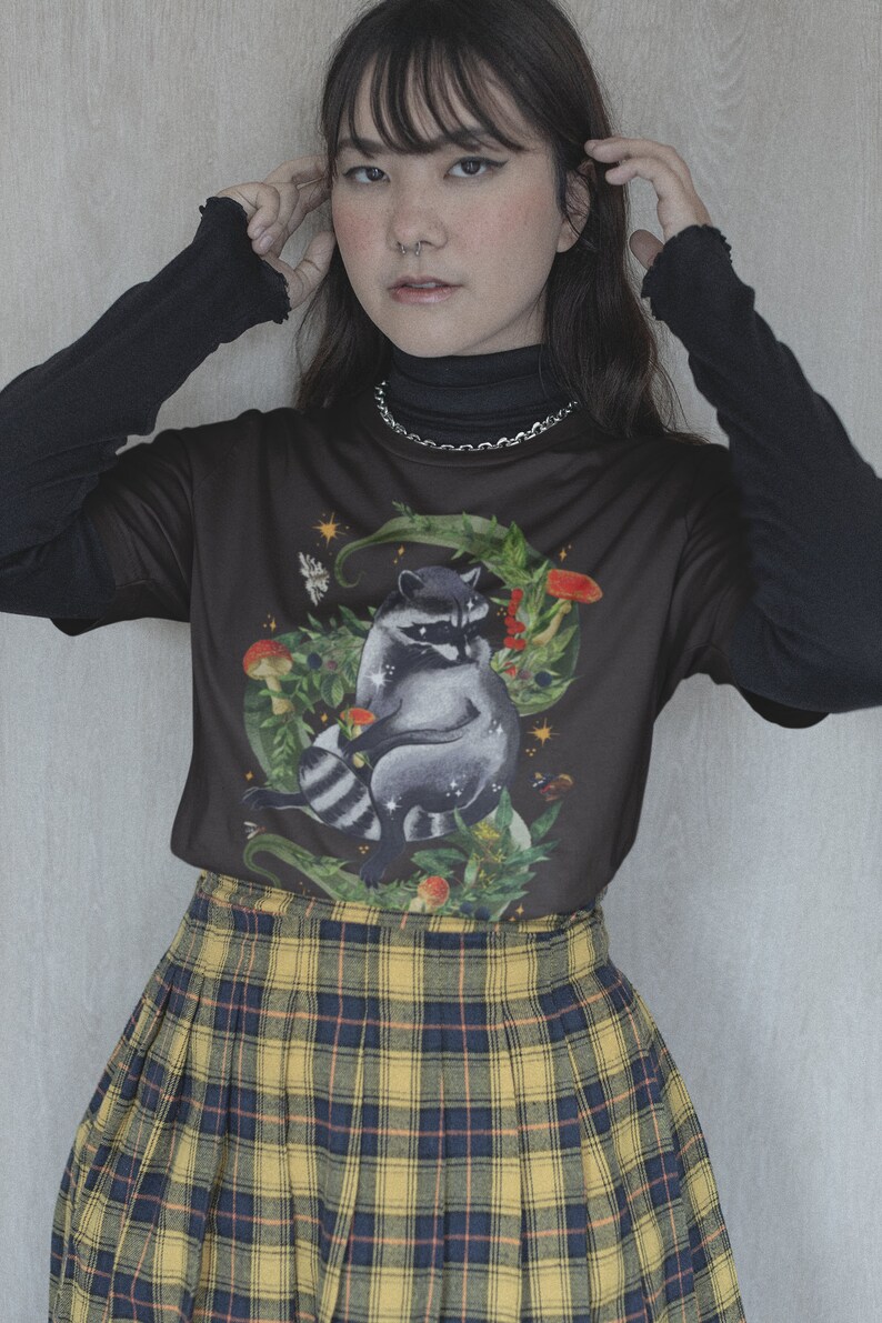 May include: A black t-shirt with a raccoon illustration surrounded by leaves, mushrooms, and stars. The shirt is worn over a black turtleneck and paired with a yellow and black plaid skirt.