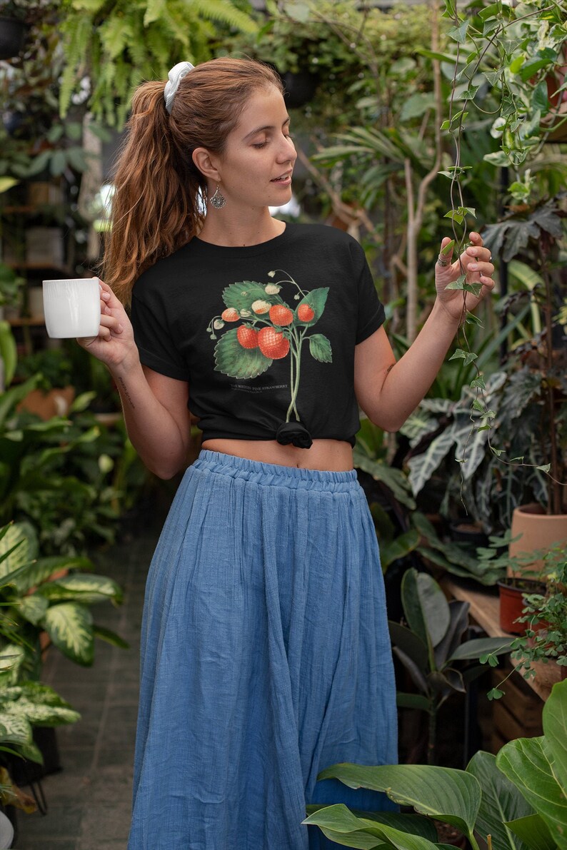 Peut inclure: Une femme portant un t-shirt noir avec une illustration vintage de fraises. Elle tient une tasse blanche dans une main et touche une vigne verte de l'autre. Elle porte une jupe bleue et se tient dans une serre.