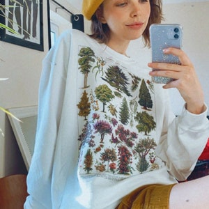Puede incluir: Una sudadera blanca con un estampado bot&aacute;nico vintage de &aacute;rboles y flores. La sudadera se lleva con una boina amarilla mostaza y unos pantalones cortos de pana amarilla mostaza.