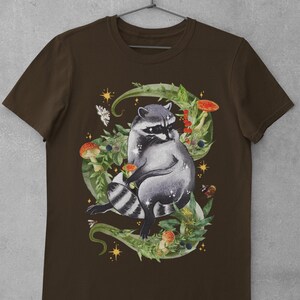 May include: A brown t-shirt with a graphic of a raccoon sitting in a bed of green leaves, mushrooms, and berries. The raccoon is surrounded by gold stars and a bee.