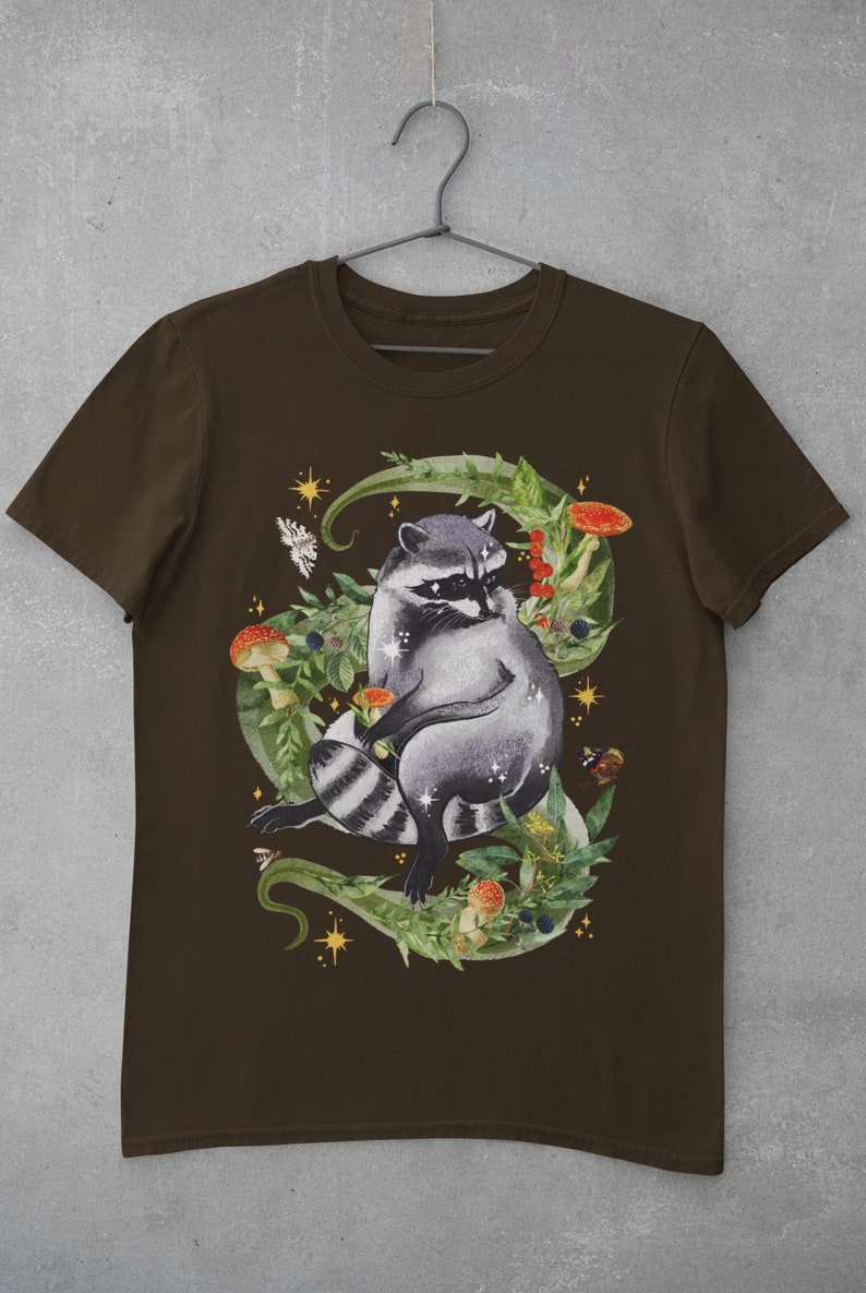 May include: A brown t-shirt with a graphic of a raccoon sitting in a bed of green leaves, mushrooms, and berries. The raccoon is surrounded by gold stars and a bee.