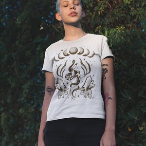 Pu&ograve; includere: Maglietta bianca con una stampa grafica di un serpente avvolto attorno a cristalli e un design a mezzaluna. La grafica &egrave; in marrone e nero.