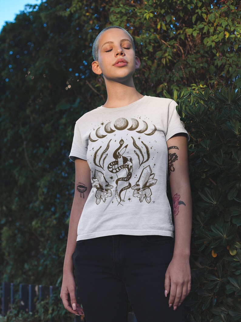 Pu&ograve; includere: Maglietta bianca con una stampa grafica di un serpente avvolto attorno a cristalli e un design a mezzaluna. La grafica &egrave; in marrone e nero.