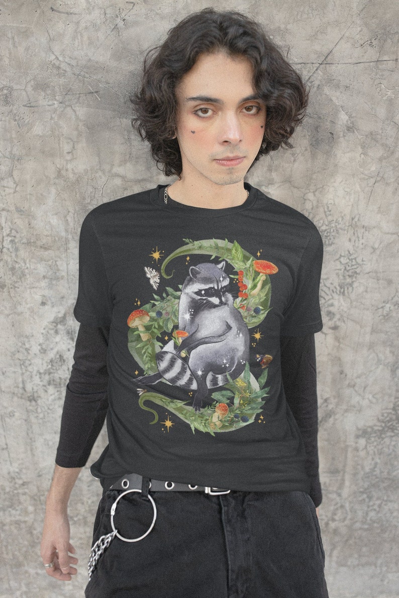 May include: Black long-sleeve t-shirt with a graphic print of a raccoon sitting in a circle of leaves and mushrooms. The raccoon has a black mask and white stripes on its tail. The design is surrounded by stars and other foliage.