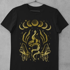 Pu&ograve; includere: Maglietta nera con un design dorato che presenta un serpente avvolto attorno a una mano, cristalli e lune crescenti.