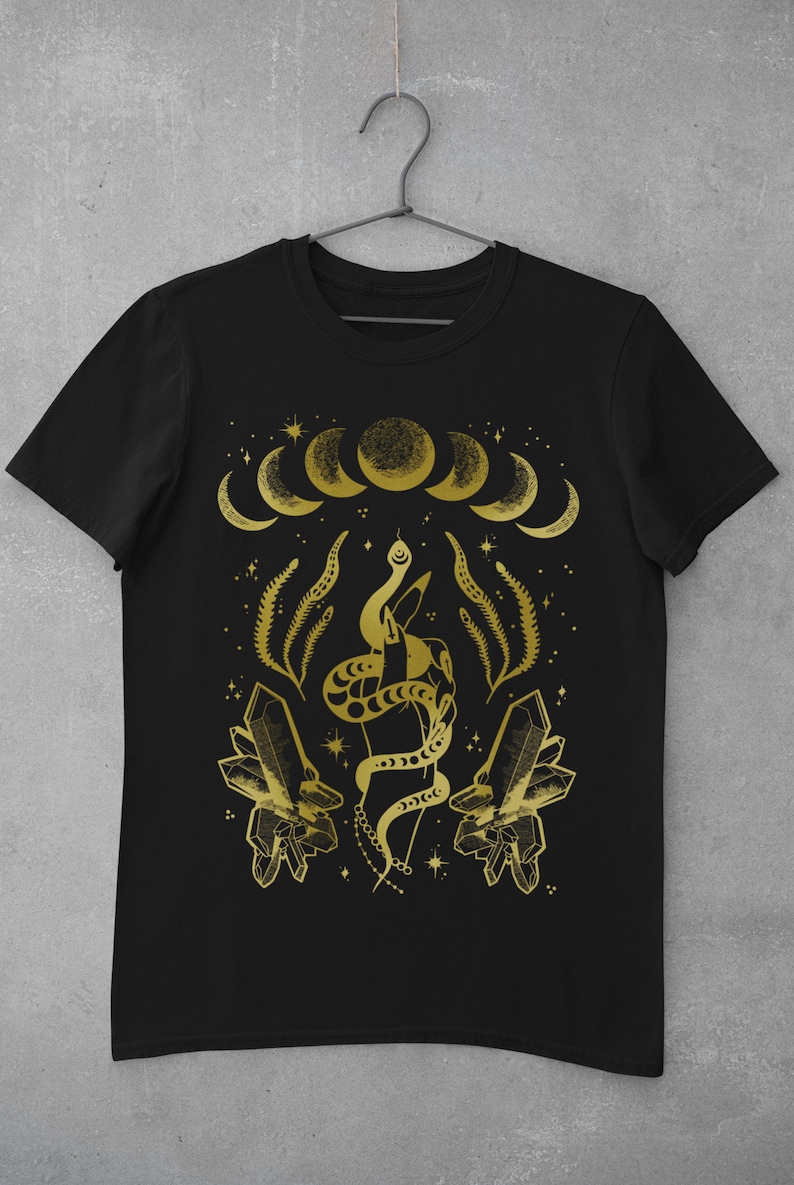 Pu&ograve; includere: Maglietta nera con un design dorato che presenta un serpente avvolto attorno a una mano, cristalli e lune crescenti.