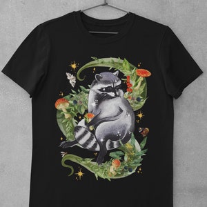 May include: Black t-shirt with a graphic of a raccoon sitting in a bed of green leaves, mushrooms, and berries. The raccoon is surrounded by gold stars and butterflies.