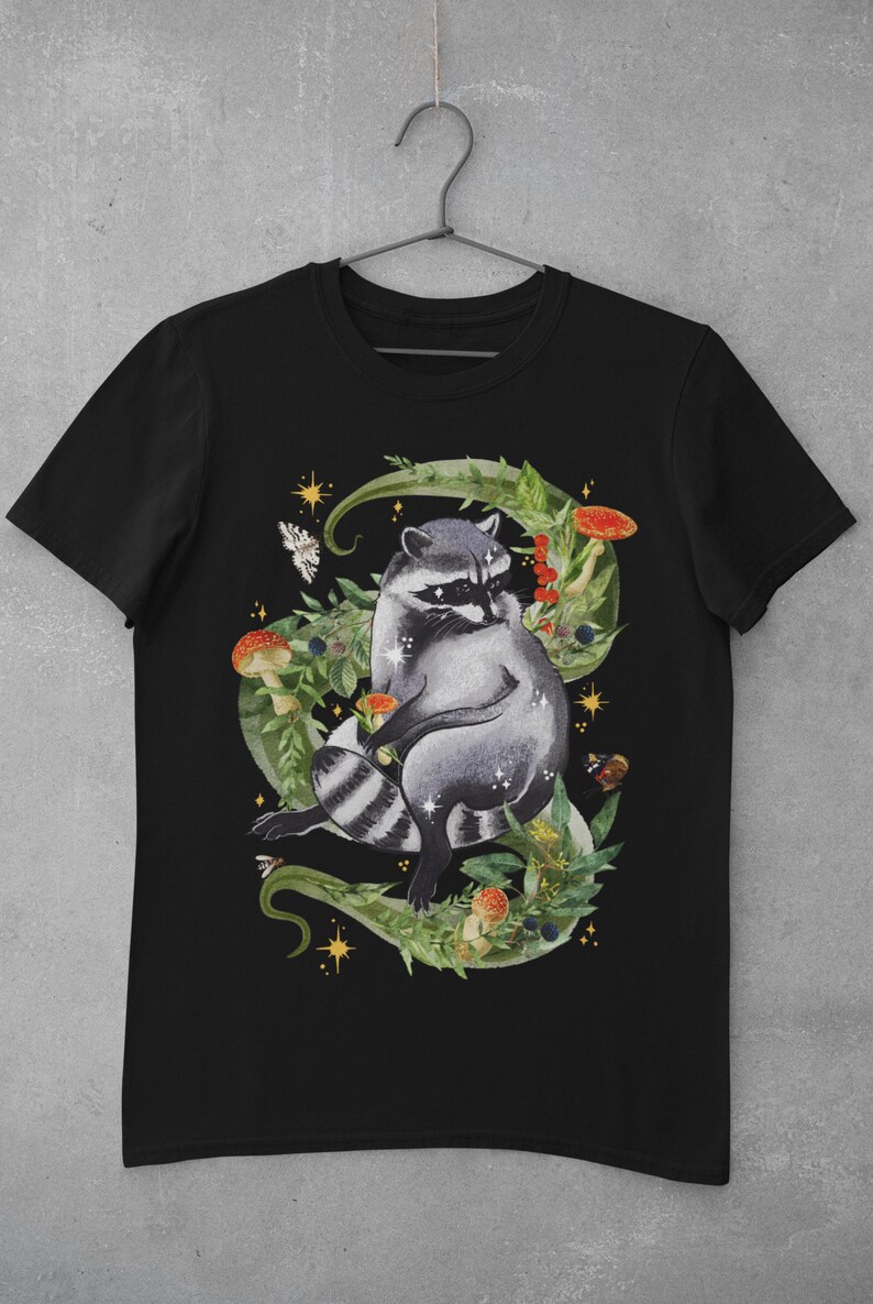 May include: Black t-shirt with a graphic of a raccoon sitting in a bed of green leaves, mushrooms, and berries. The raccoon is surrounded by gold stars and butterflies.