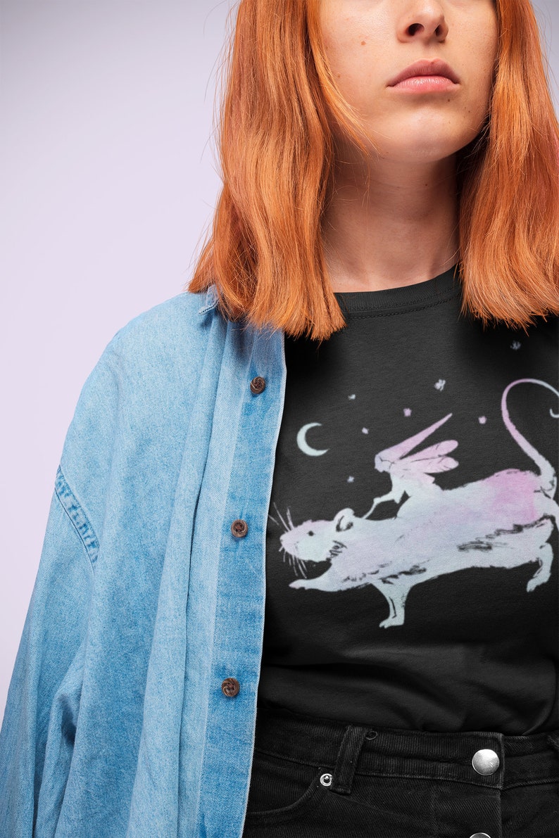 Puede incluir: Una camiseta negra con un gr&aacute;fico de una rata blanca con un hada montada en su espalda. El hada tiene alas y sostiene una varita m&aacute;gica. El fondo es azul oscuro con estrellas blancas.