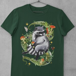 May include: A dark green t-shirt with a raccoon surrounded by leaves, mushrooms, and berries. The raccoon is sitting on a vine with its tail curled around its body. The design is in a watercolor style.