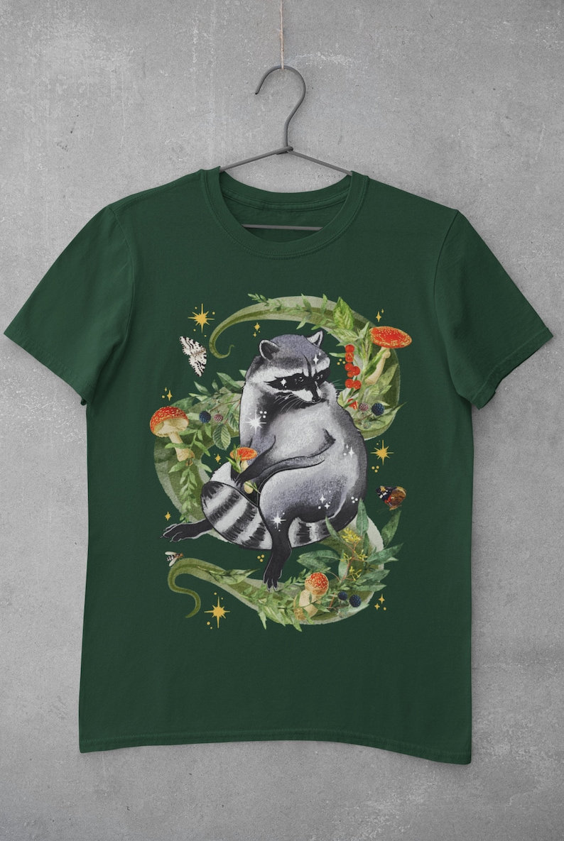 May include: A dark green t-shirt with a raccoon surrounded by leaves, mushrooms, and berries. The raccoon is sitting on a vine with its tail curled around its body. The design is in a watercolor style.