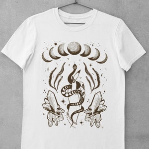 Pu&ograve; includere: Maglietta bianca con un design grafico marrone che rappresenta una mano che tiene un serpente, cristalli e una luna crescente.