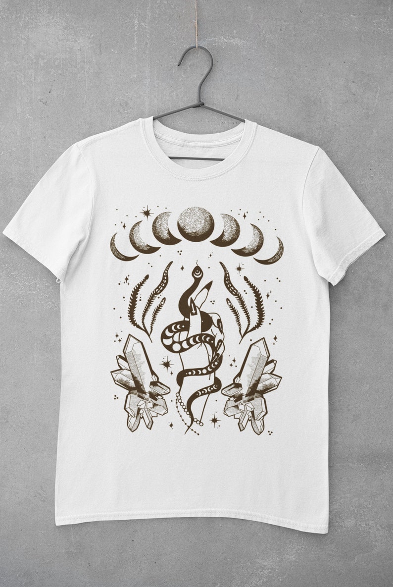 Pu&ograve; includere: Maglietta bianca con un design grafico marrone che rappresenta una mano che tiene un serpente, cristalli e una luna crescente.