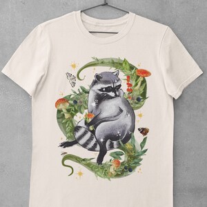 May include: A white t-shirt with a graphic of a raccoon sitting in a circle of green leaves, mushrooms, and berries. The raccoon is gray and black with a white mask around its eyes. There are small gold stars and butterflies around the raccoon.