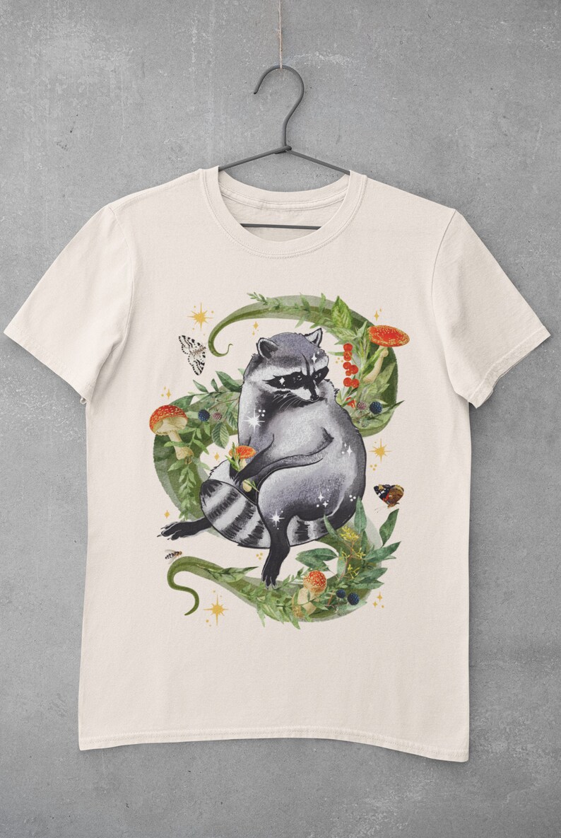 May include: A white t-shirt with a graphic of a raccoon sitting in a circle of green leaves, mushrooms, and berries. The raccoon is gray and black with a white mask around its eyes. There are small gold stars and butterflies around the raccoon.
