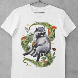 May include: White t-shirt with a watercolor illustration of a raccoon sitting in a circle of green vines, leaves, and mushrooms. The raccoon is gray with black stripes and a black mask. There are also butterflies and stars in the design.