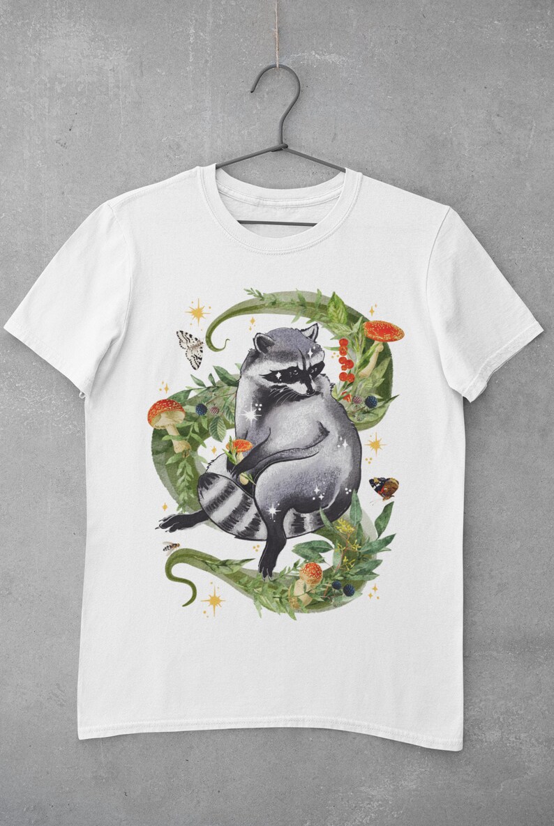 May include: White t-shirt with a watercolor illustration of a raccoon sitting in a circle of green vines, leaves, and mushrooms. The raccoon is gray with black stripes and a black mask. There are also butterflies and stars in the design.
