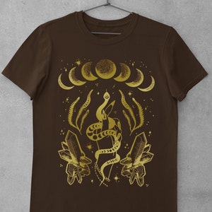 Pu&ograve; includere: Una maglietta marrone con un design dorato che presenta un serpente avvolto attorno a una mano, cristalli e una luna crescente.