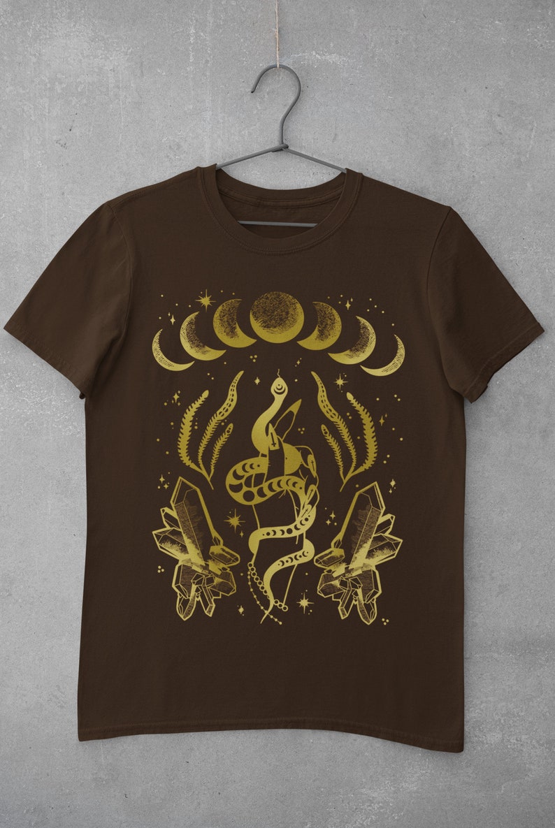 Pu&ograve; includere: Una maglietta marrone con un design dorato che presenta un serpente avvolto attorno a una mano, cristalli e una luna crescente.