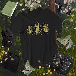 Könnte beinhalten: Schwarzes T-Shirt mit einem Aufdruck von drei Insekten, darunter ein Hirschkäfer, ein Käfer mit langem Rüssel und ein Käfer mit rundem Körper. Die Insekten sind in einer Reihe angeordnet und in Gold umrandet.