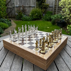 May include: A wooden chessboard with a set of silver and gold chess pieces. The board is set up for a game of chess, with the pieces arranged in their starting positions.