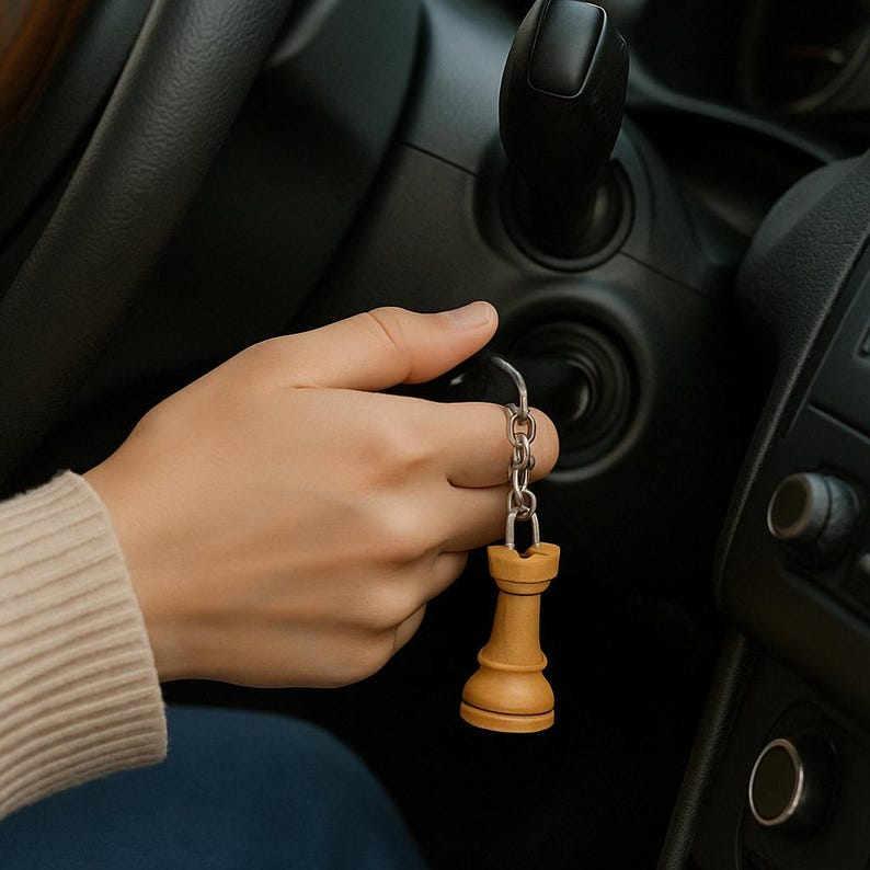 May include: A beige chess rook keychain hanging from a silver chain. The keychain is held in a hand near a car's ignition. The rook is a light brown color with a darker brown base.