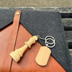 May include: A wooden chess king keychain and a wooden tag with a silver keyring. The chess piece is light brown and detailed. The tag is rectangular with rounded edges. The items rest on a leather and felt surface.