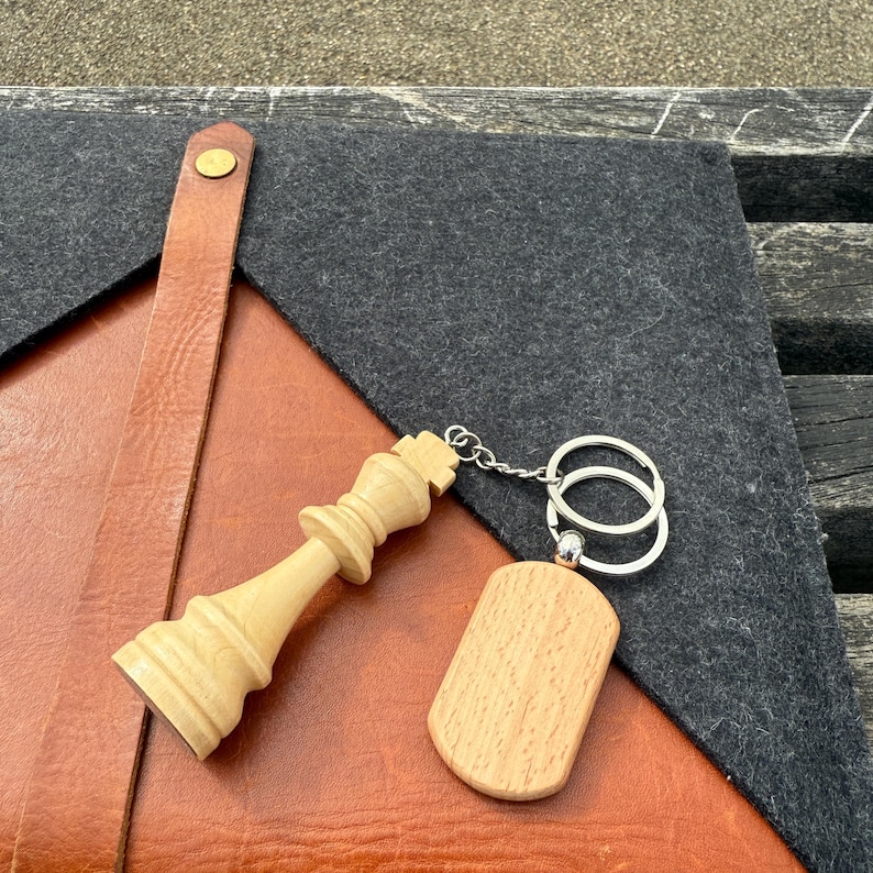 May include: A wooden chess king keychain and a wooden tag with a silver keyring. The chess piece is light brown and detailed. The tag is rectangular with rounded edges. The items rest on a leather and felt surface.