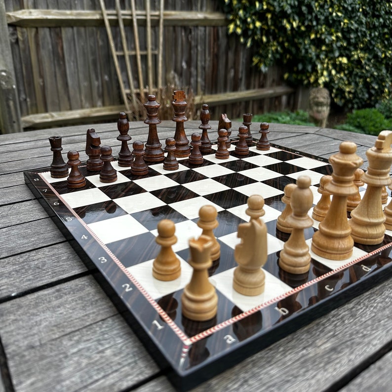 May include: A black and white chessboard with wooden chess pieces. The white pieces are on the right side of the board and the black pieces are on the left side. The board is set up for a game of chess.