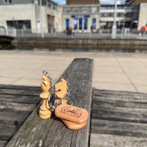 May include: Three wooden chess piece keychains: a bishop, a knight, and a rectangular piece with a chess-themed engraving. The keychains are made of light-colored wood and are attached to silver keyrings. The pieces are displayed on a weathered wooden bench.