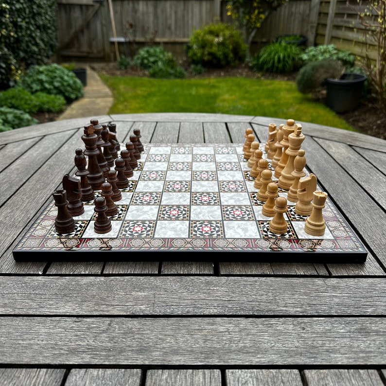 May include: A wooden chessboard with black and white squares, featuring a decorative inlay design. The board is set up with black and white chess pieces, with the white pieces on the right side and the black pieces on the left side.