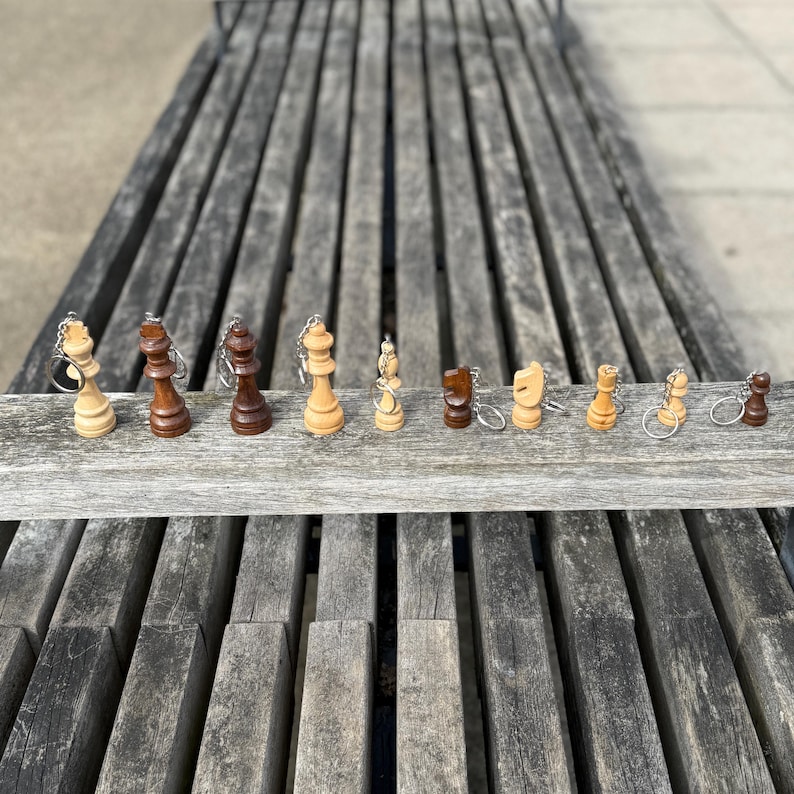 May include: A collection of miniature chess piece keychains in various colors, including light and dark wood tones. The pieces are arranged on a wooden plank, showcasing the king, queen, bishop, knight, rook, and pawn. The keychains are attached with silver metal rings.