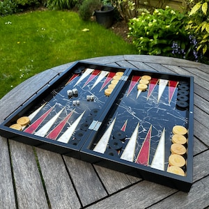 May include: A black and white backgammon board with red and white stripes. The board is open and shows black playing pieces and white playing pieces. The board is made of wood and has a marble-like design.