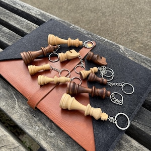 May include: A collection of wooden chess piece keychains in various colors, including light and dark brown. The keychains are attached to silver rings and chains, resting on a brown leather and gray felt surface.
