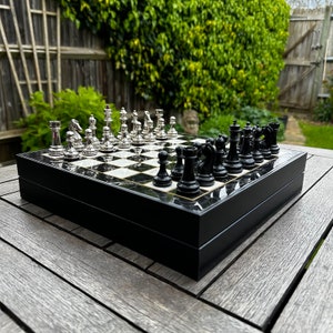 May include: A black and white chessboard with silver chess pieces on one side and black chess pieces on the other side. The chessboard is in a black wooden box.