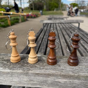 May include: Four wooden chess piece keychains. Two are light-colored, and two are dark brown. Each keychain features a chess piece design, including a queen and a king, and a silver-colored chain and ring.