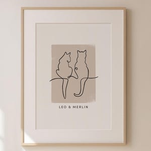 Könnte beinhalten: Ein minimalistischer Line-Art-Druck von zwei Katzen, die Rücken an Rücken sitzen, mit dem Text "Leo & Merlin" darunter. Der Druck ist auf beigefarbenem Hintergrund und in einem weißen Rahmen gerahmt.
