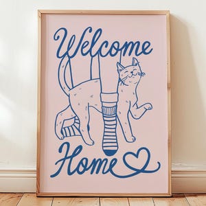 May include: A framed print with a light pink background and a blue illustration. The illustration features a cat, a pair of legs with a striped sock, and the words "Welcome Home" in a script font. A heart symbol is next to the word "Home".