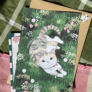 May include: A colorful illustration of a cat named Mabel lying in a field of green grass and white flowers. The cat has brown, white, and gray fur and is wearing a pink collar. The illustration is surrounded by a brown paper envelope.