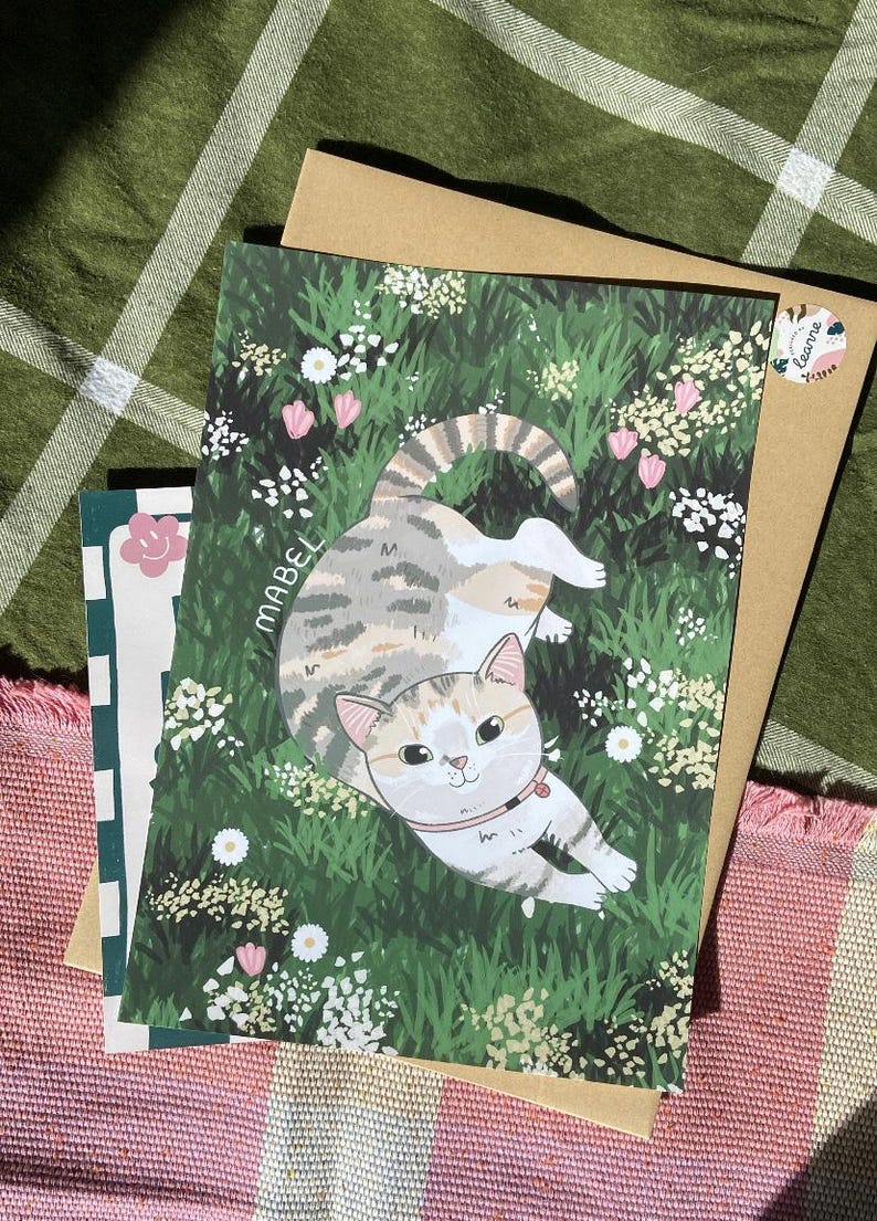 May include: A colorful illustration of a cat named Mabel lying in a field of green grass and white flowers. The cat has brown, white, and gray fur and is wearing a pink collar. The illustration is surrounded by a brown paper envelope.
