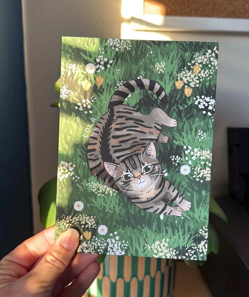 May include: A gray tabby cat with green eyes lies on its back in a field of white flowers and green grass. The cat's name, "LOLA", is written in white letters on the grass.
