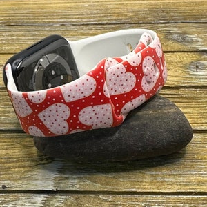 May include: A black smartwatch sits in a white and red heart-patterned strap. The strap, decorated with pink polka dots, rests on a dark grey stone, against a wooden backdrop. This accessory blends technology with a romantic design.