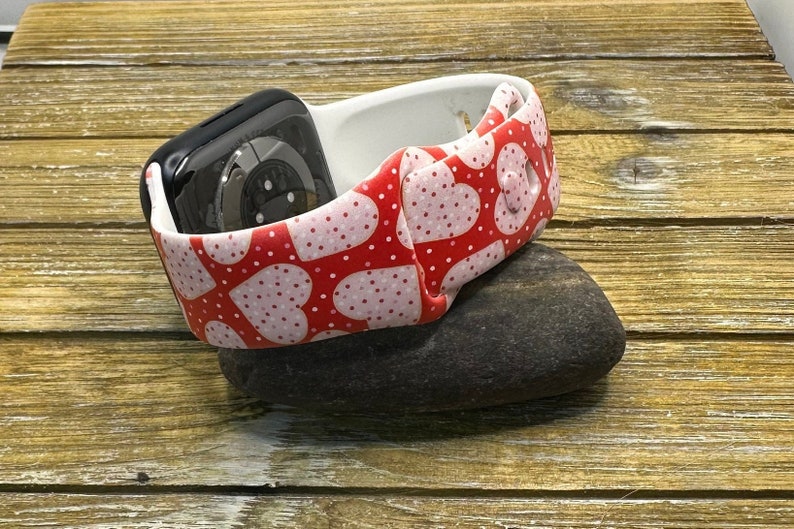 May include: A black smartwatch sits in a white and red heart-patterned strap. The strap, decorated with pink polka dots, rests on a dark grey stone, against a wooden backdrop. This accessory blends technology with a romantic design.