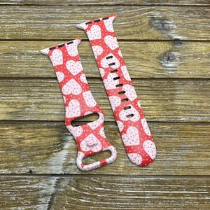 May include: Red and white Apple Watch strap with a heart pattern. The strap features a repeating design of white hearts and small white dots on a red background. The strap is made of a flexible material and has multiple holes for adjustment.