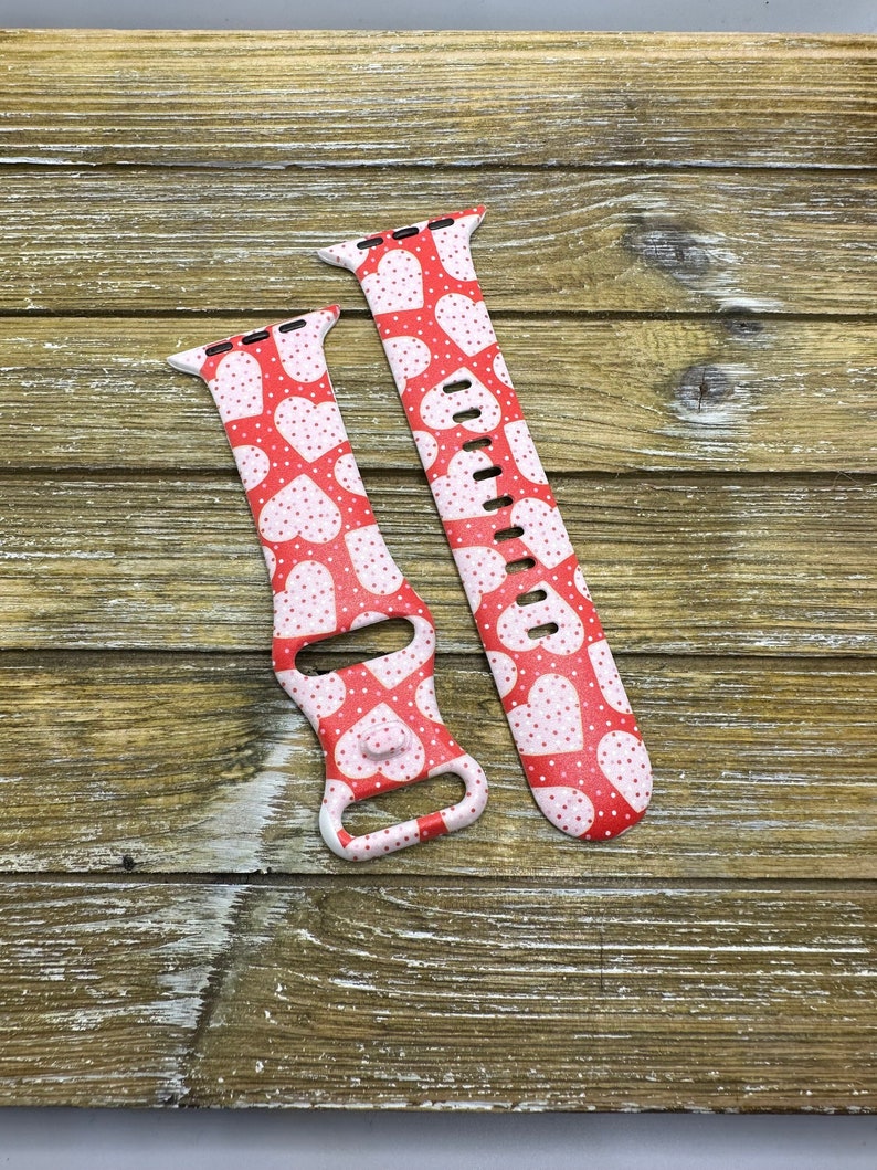 May include: Red and white Apple Watch strap with a heart pattern. The strap features a repeating design of white hearts and small white dots on a red background. The strap is made of a flexible material and has multiple holes for adjustment.