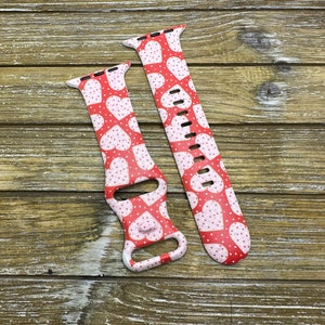 May include: Two red and white patterned watch straps. The straps feature a repeating pattern of white hearts and small white dots on a red background. The straps are designed to fit a smartwatch and are displayed on a wooden surface.