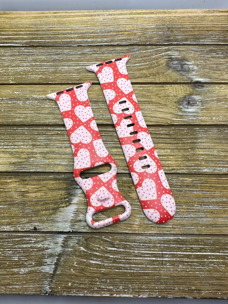 May include: Two red and white patterned watch straps. The straps feature a repeating pattern of white hearts and small white dots on a red background. The straps are designed to fit a smartwatch and are displayed on a wooden surface.